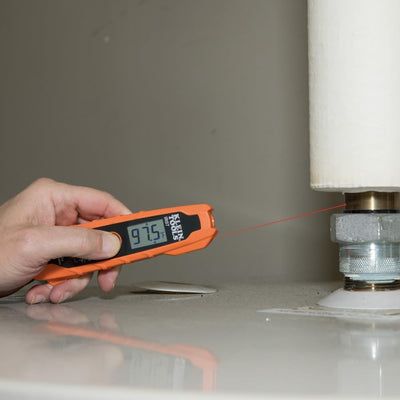 Hand holding a digital thermometer near a pipe with a red laser pointer.