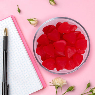 Red heart-shaped candies in a glass bowl on a pink surface with a pen and notebook.